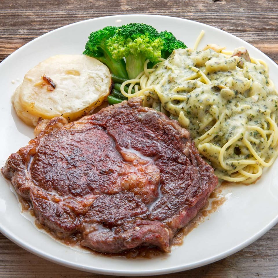 Grilled steak, creamy pasta, roasted potatoes, and broccoli on a white plate at Delicious Food Corner.