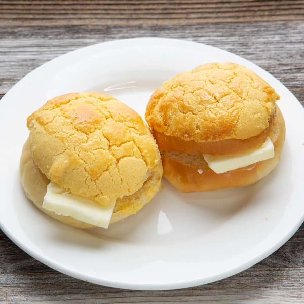 Two pineapple buns with butter slices on a white plate, against a rustic wooden background.